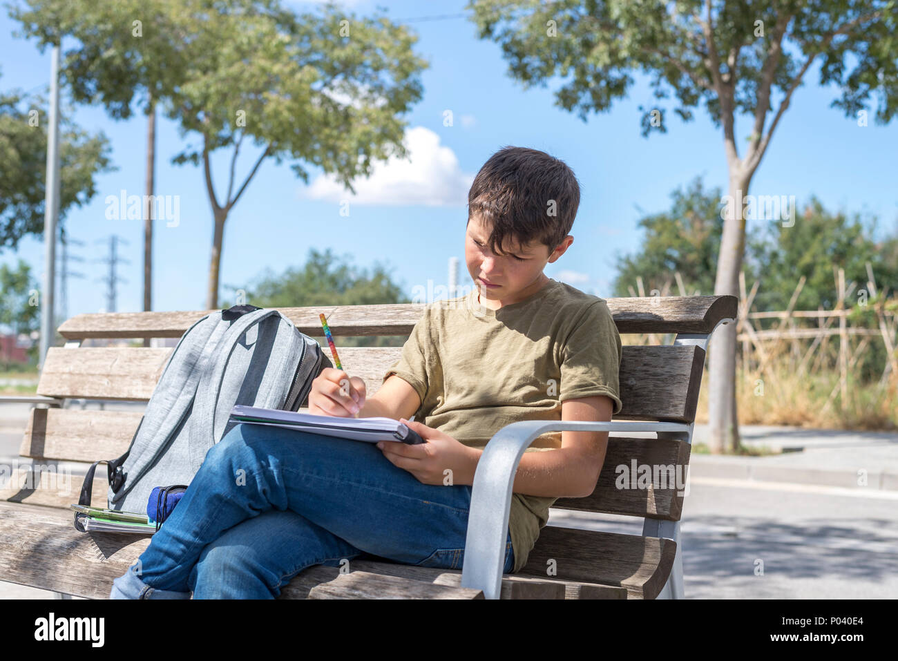 Portrait of a teenager boy sitting on a wood park bench doing his college homework and studying writing notes during a sunny day. Outdoors student lif Stock Photo