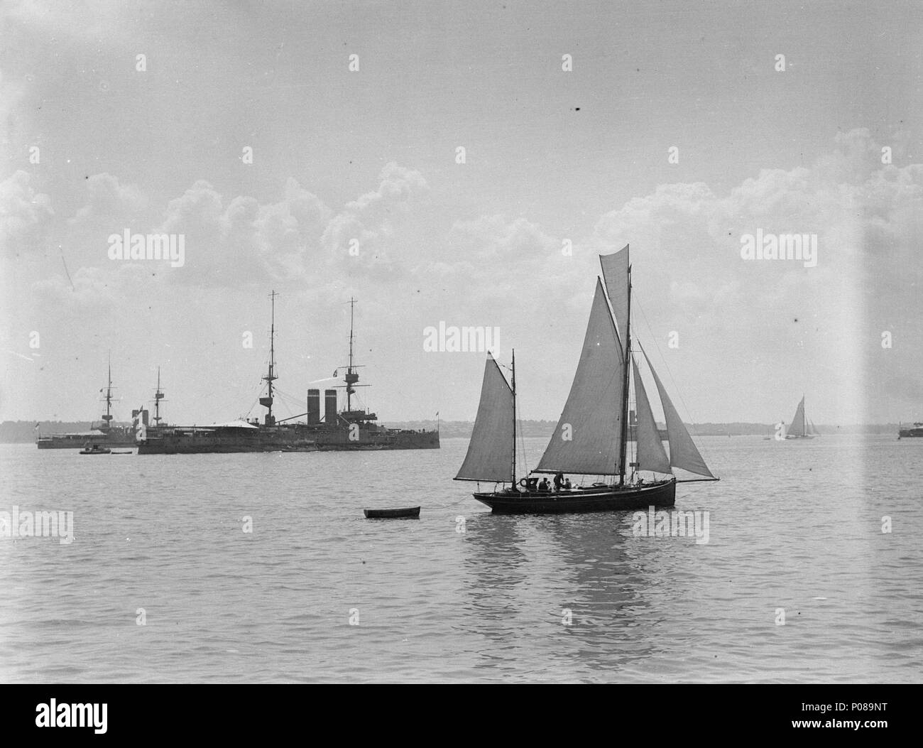 .  English: A general view of the ships at Spithead during the Fleet Review A general view looking towards the Isle of Wight of the line of warships for the Naval Fleet Review at Spithead. In the foreground is an unnamed ketch-rigged yacht under sail, towing its tender. In the middle distance is the battleship 'Dominion' (1903), partially obscuring the battleship 'Albion' (1898). To the right of the photograph can be seen the stern of the battleship 'Formidable' (1898). The coast of the Isle of Wight is in the background. A general view of the ships at Spithead during the Fleet Review  . 12 Ju Stock Photo