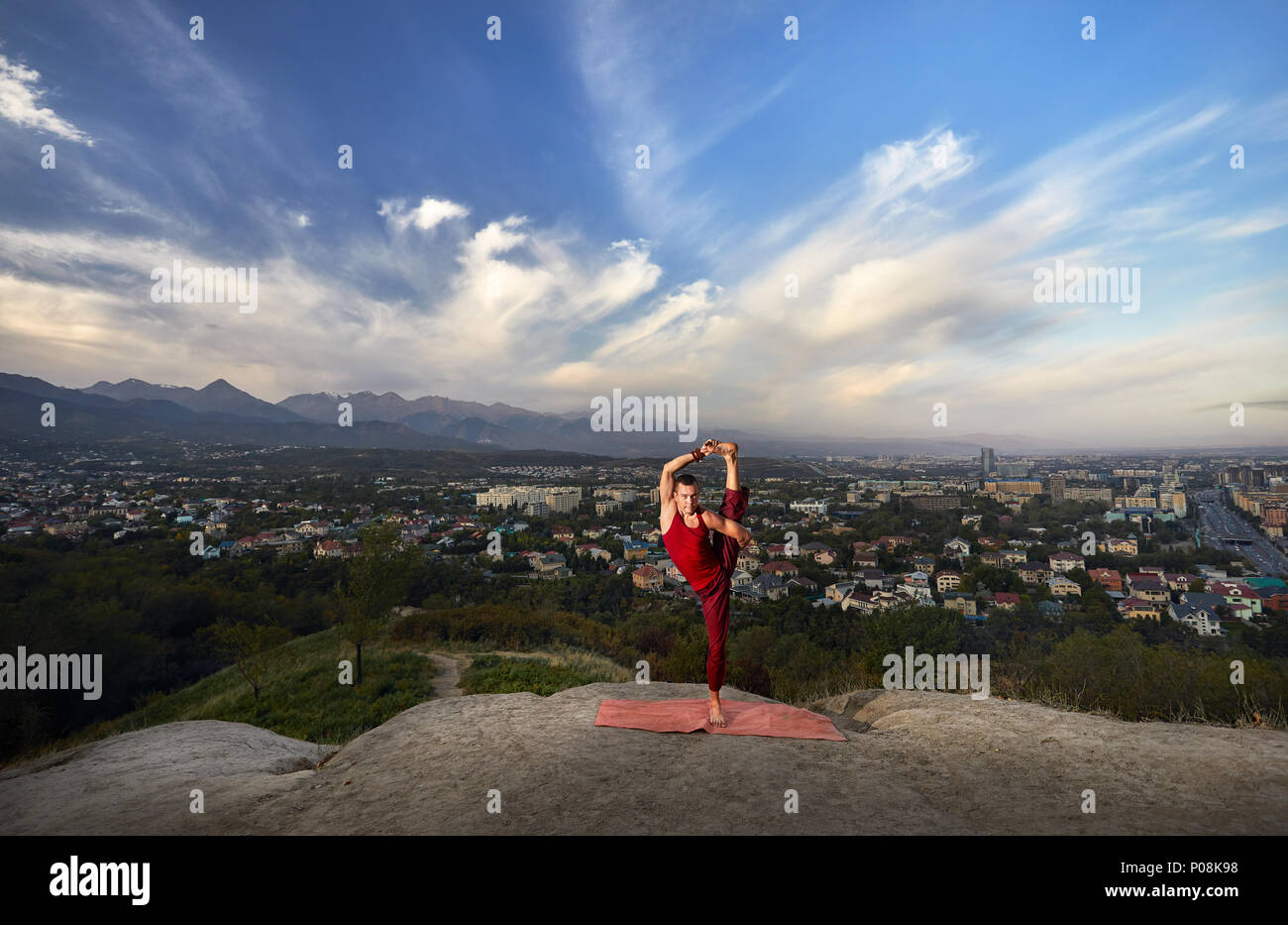 Fit man in red costume doing yoga balance asana in the park with city and mountain on background in Almaty, Kazakhstan Stock Photo