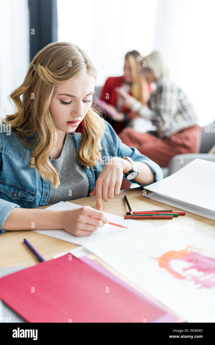 young blonde magazine editor drawing sketches in modern office, colleagues sitting behind Stock Photo