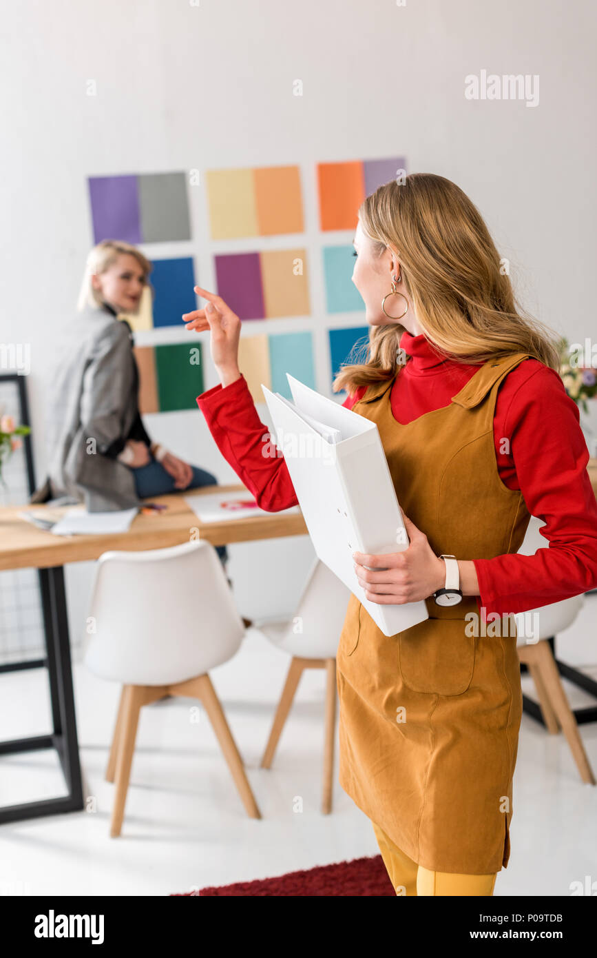 attractive magazine editor with folder pointing at colleague and color palette on wall behind Stock Photo