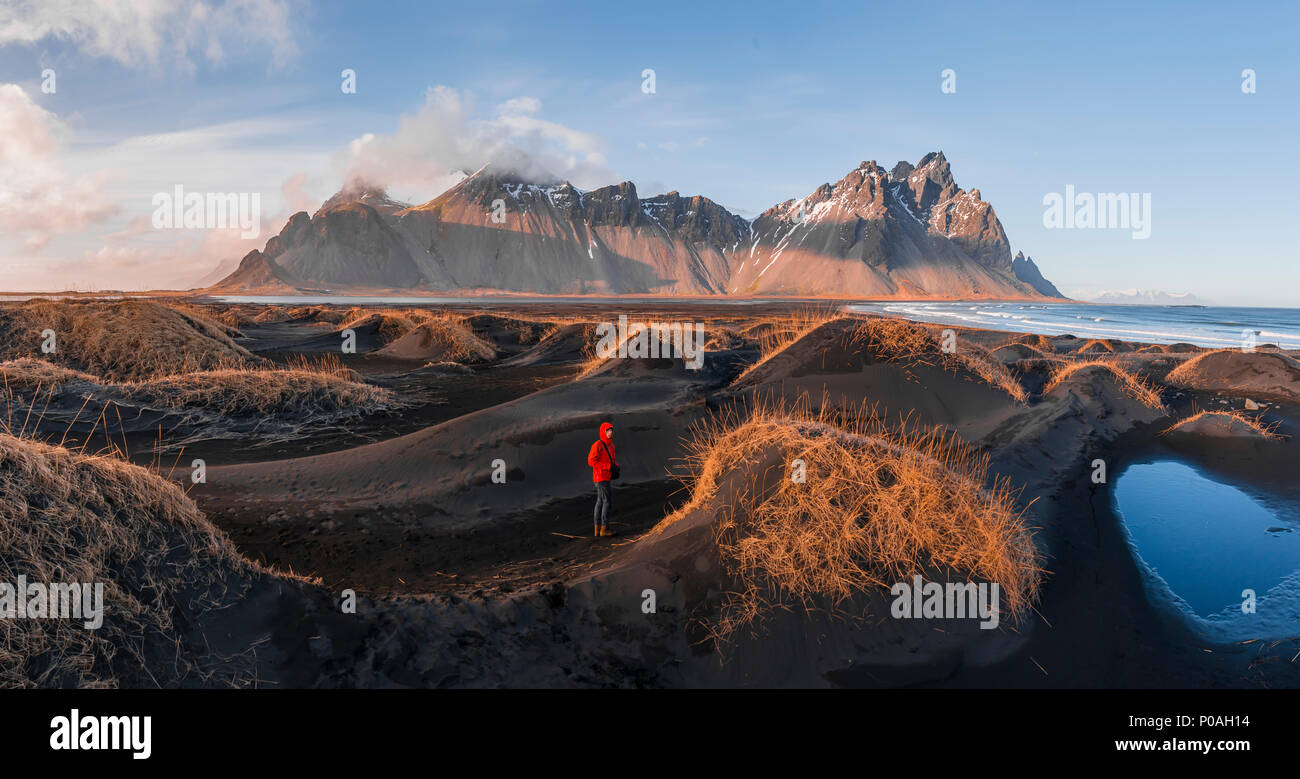 Evening atmosphere at the long lava beach, man between black sand dunes covered with dry grass, mountains Klifatindur Stock Photo