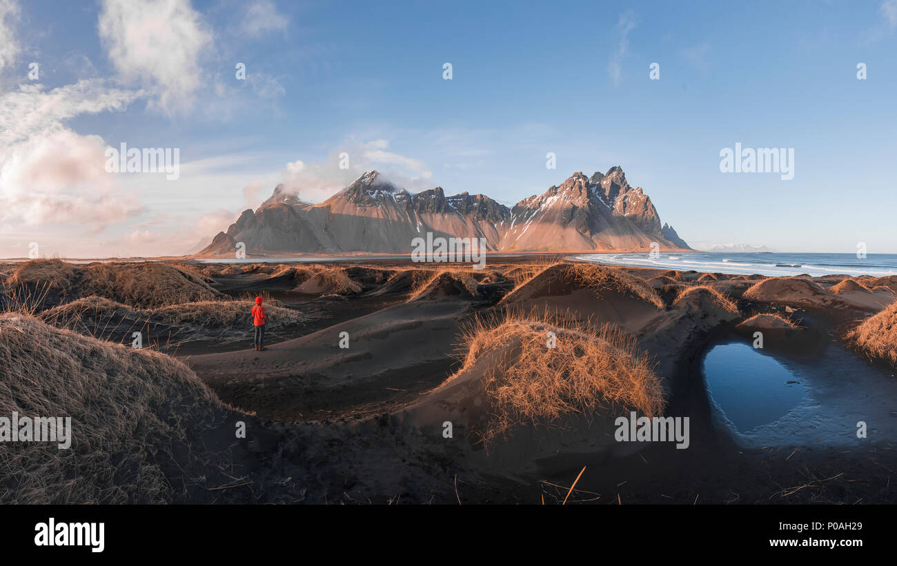 Evening atmosphere at the long lava beach, man between black sand dunes covered with dry grass, mountains Klifatindur Stock Photo