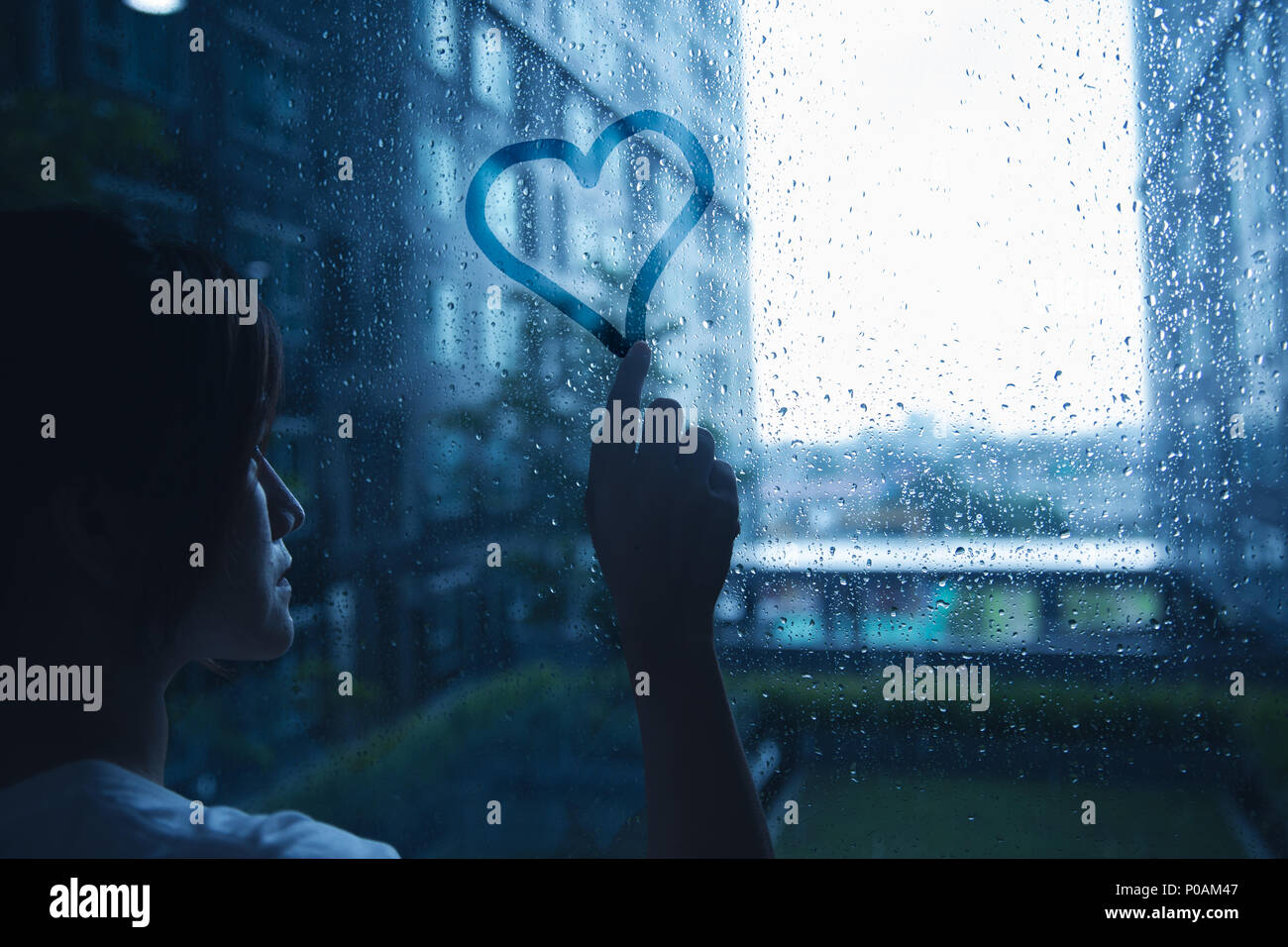 sad lonely love woman in rain draw heart on windows glass dark blue tone Stock Photo