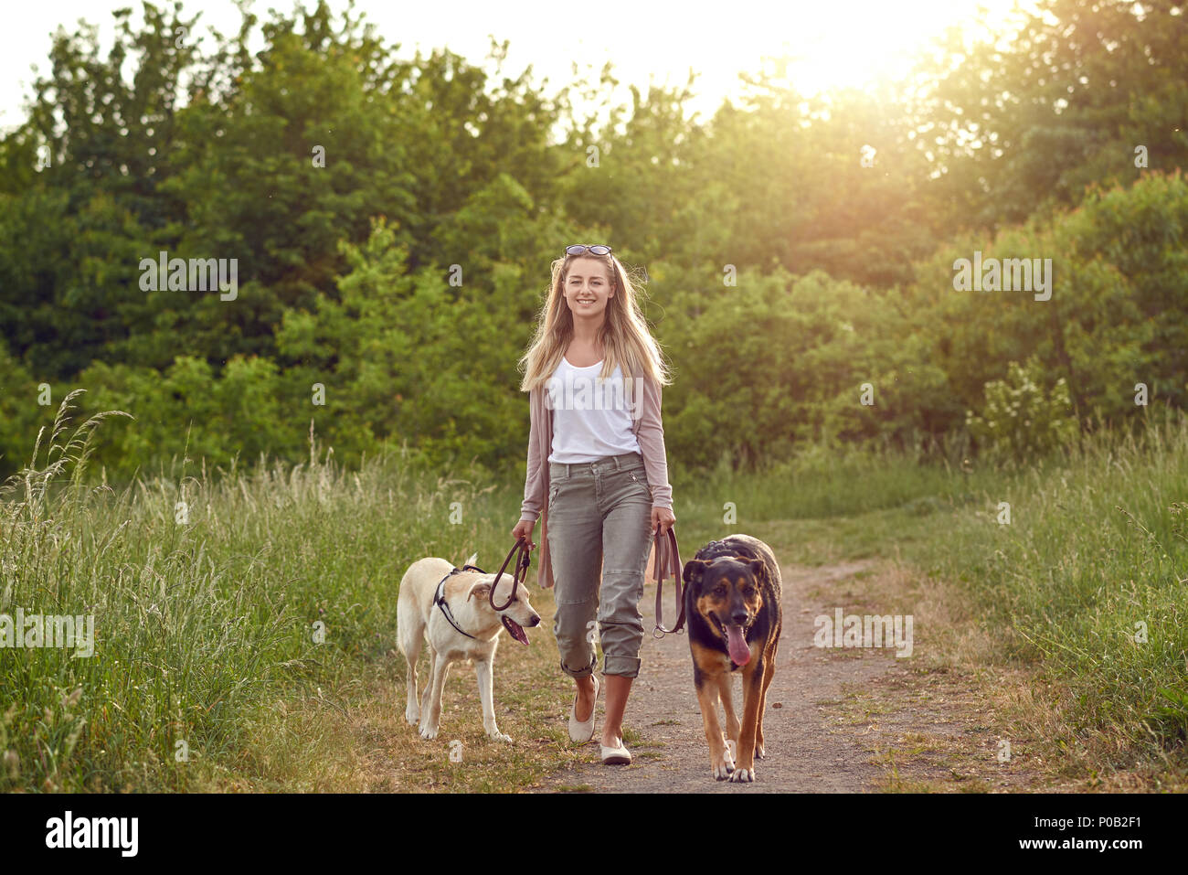 Happy young woman walking her dogs along a grassy rural track in spring looking at the camera Stock Photo