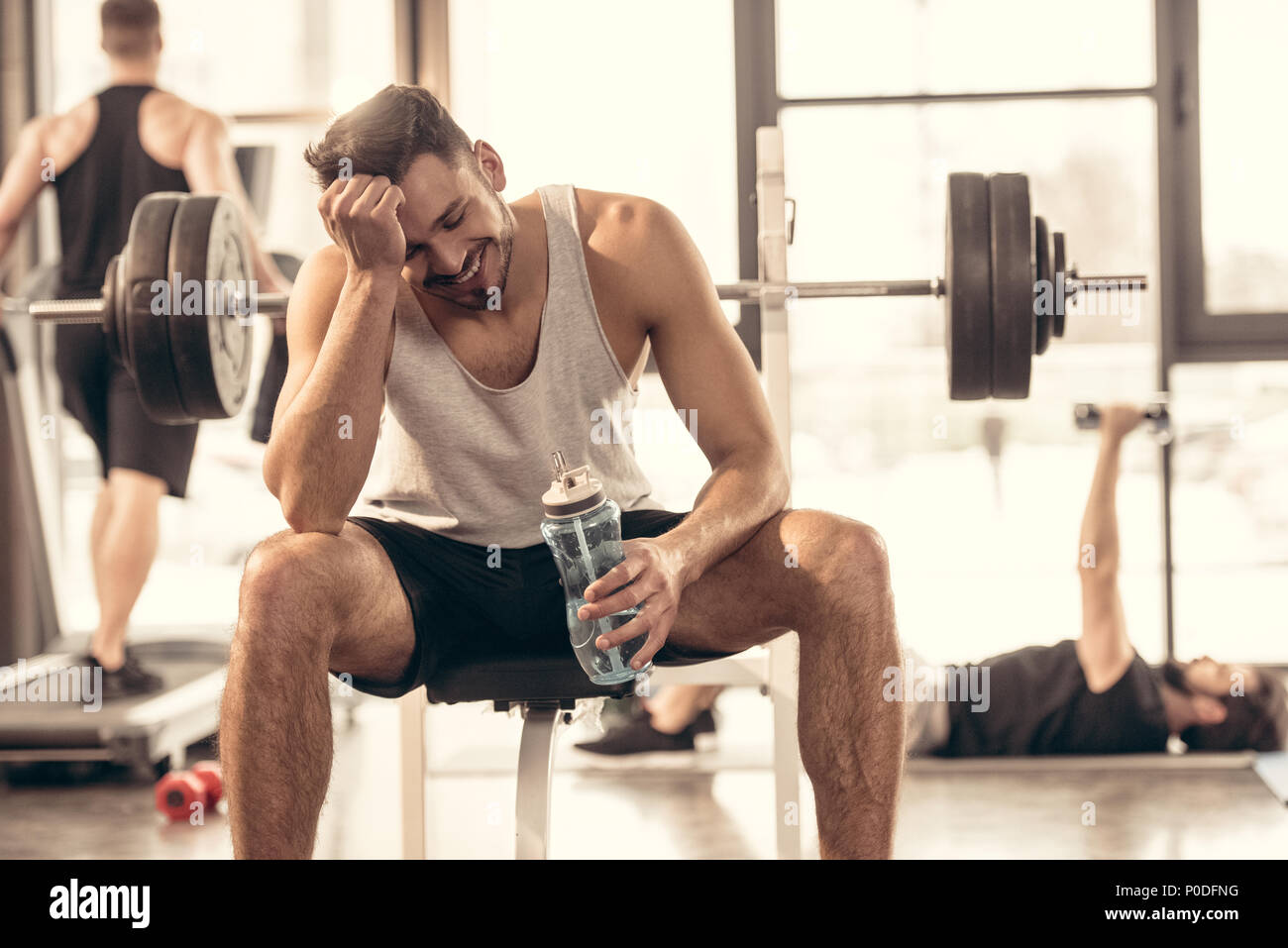 smiling sportsman sitting with bottle of water on bench press in gym Stock Photo