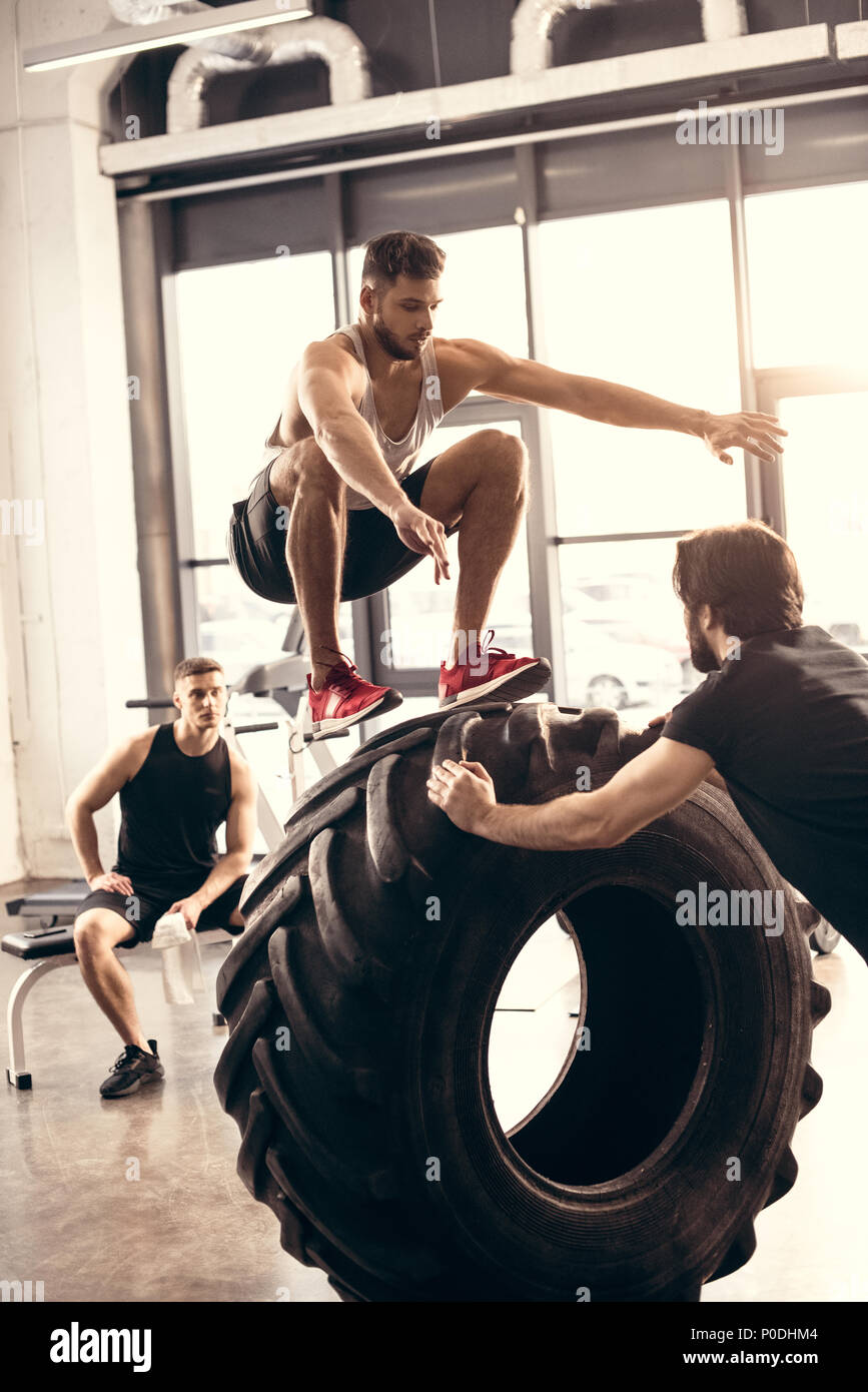 athletic young man jumping on tire while training with friends in gym Stock Photo
