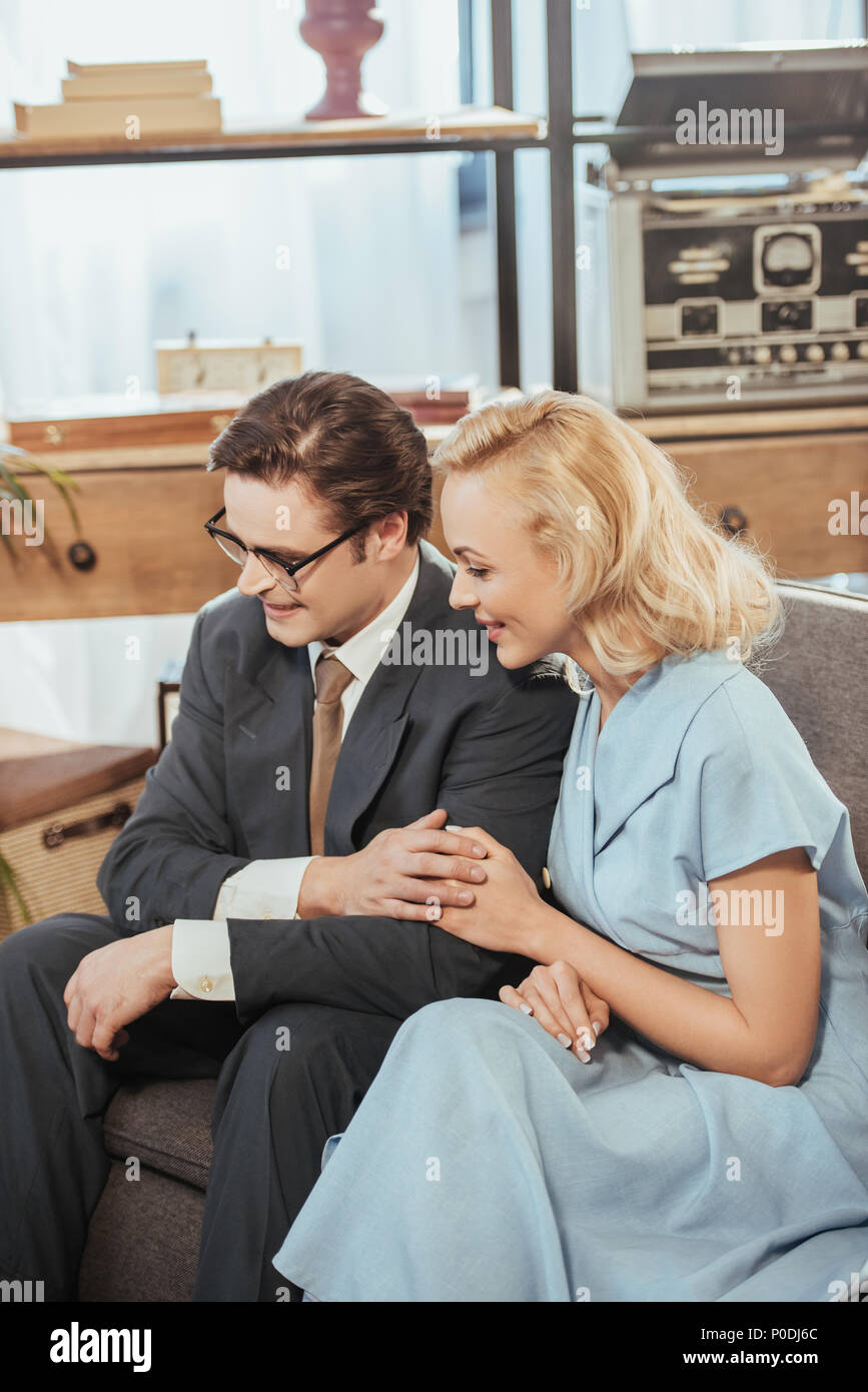 happy fifties style couple looking away while sitting on sofa and holding hands Stock Photo