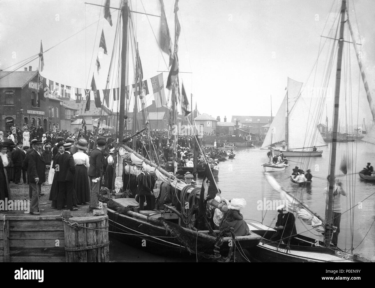 .  English: A view across Pearson's Quay along the shore of the River Colne at Rowhedge during an unidentified festival. A view looking north across Pearson's Quay along the shore of the River Colne at Rowhedge during an unidentified festival. On the left can be seen the Albion public house, with the Anchor pub in the middle distance through the rigging of various sailing craft alongside the quay. In the foreground can be seen a Colchester registered trawler, dressed overall, with visitors onboard. Cutters and rowing boats are on the water being watched by large crowds of well-dressed onlooker Stock Photo