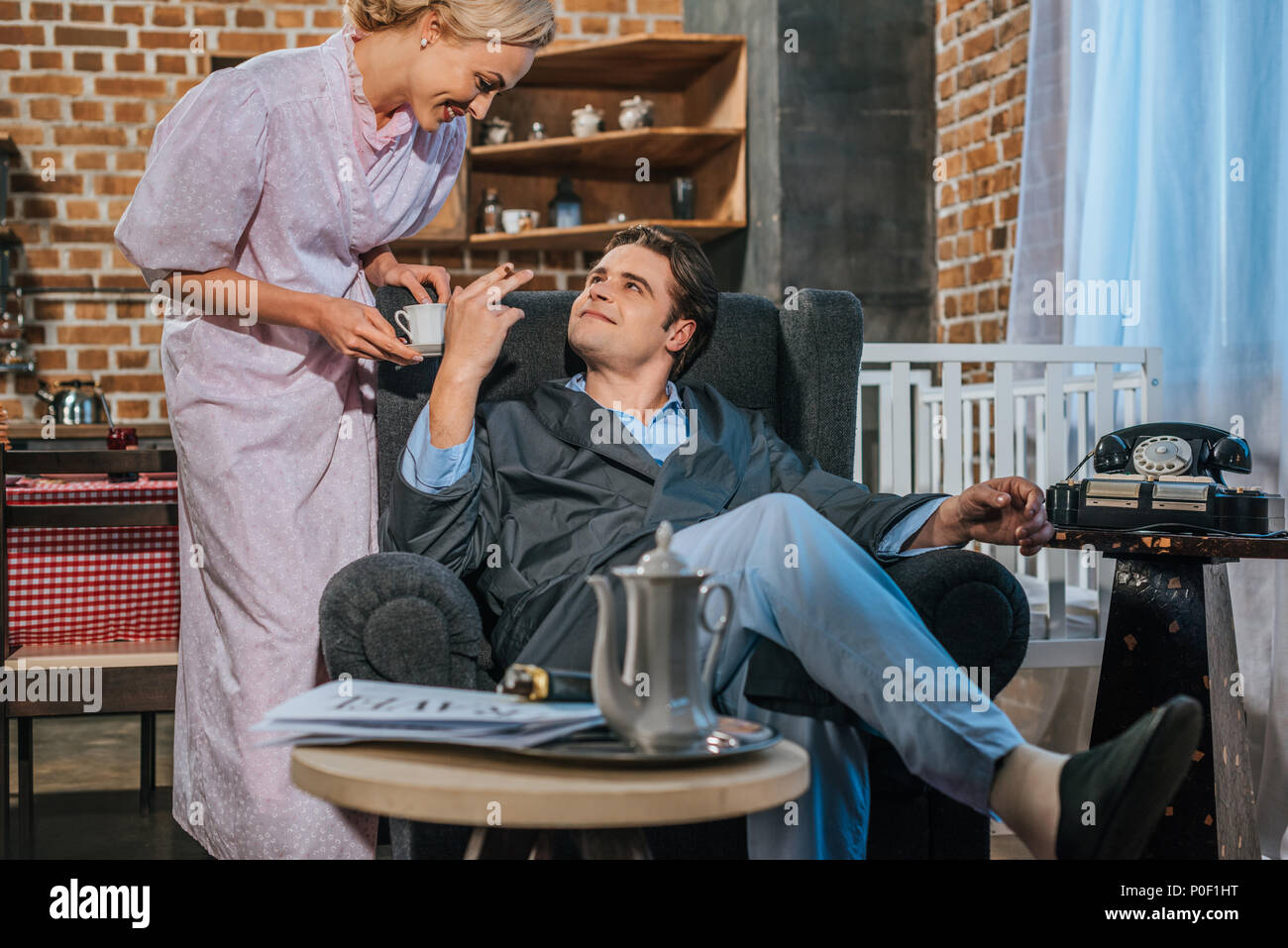 cigarette wife smiling man in robe smoking cigarette and looking at happy wife holding cup  of coffee, 1950s style Stock Photo