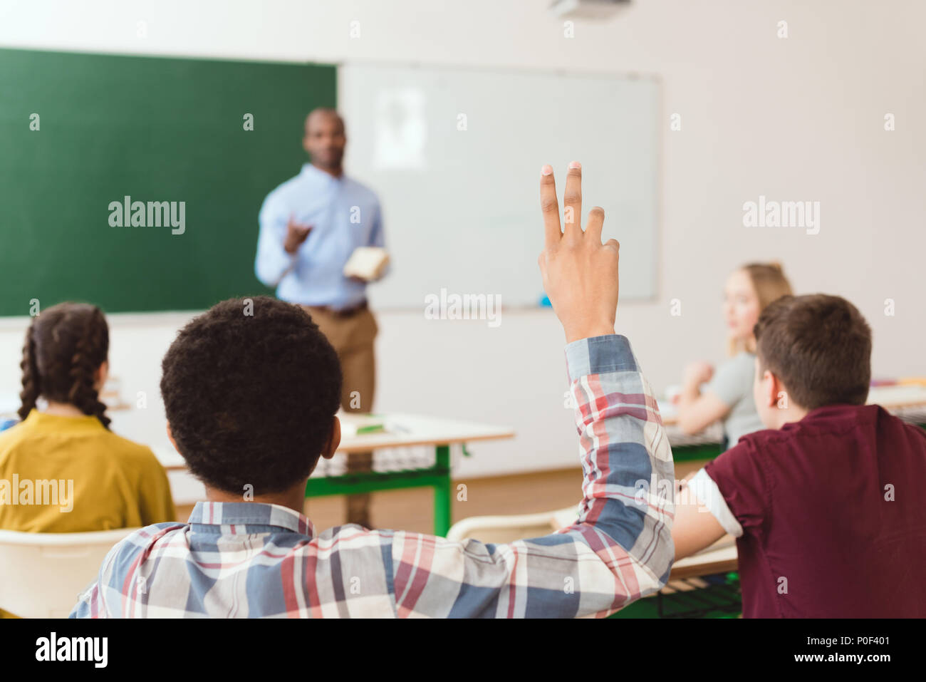 Rear view of african american boy with arm up in classroom with teacher and classmates Stock Photo