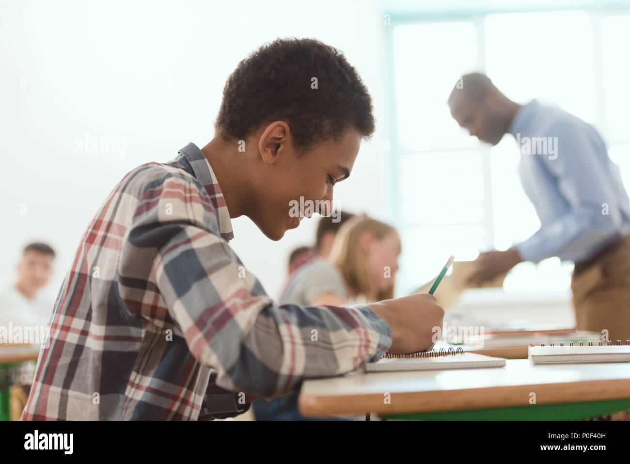 Side view of smiling african american boy writing in textbook with teacher and classmates on behind Stock Photo
