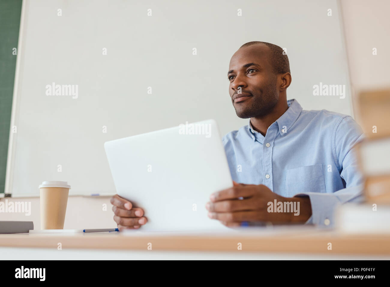 Low angle view of african american teacher sitting at desk with laptop and coffee cup Stock Photo
