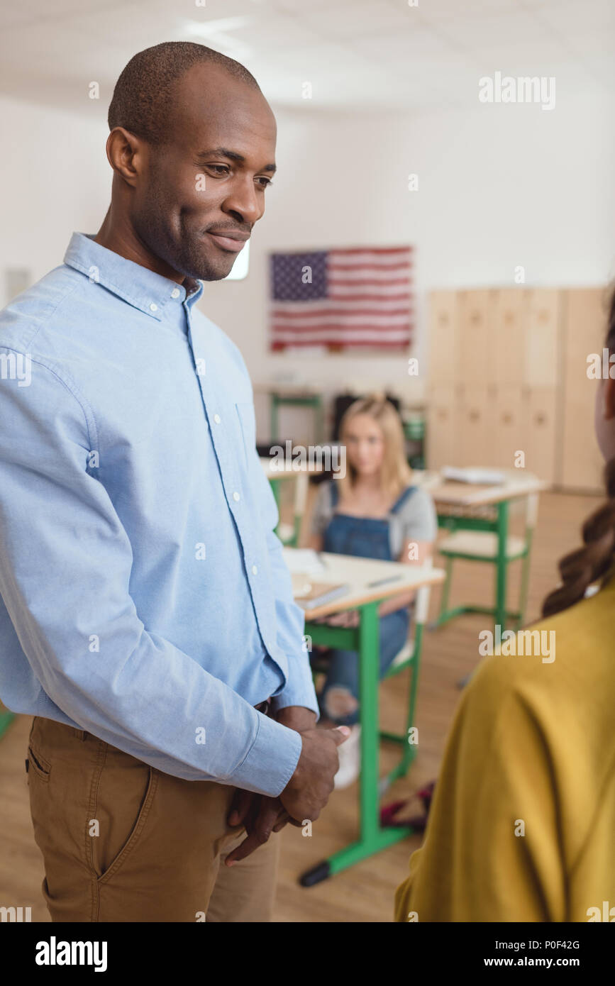Smiling african american teacher talking to schoolgirl and classmate sitting behind in classroom with flag of USA Stock Photo
