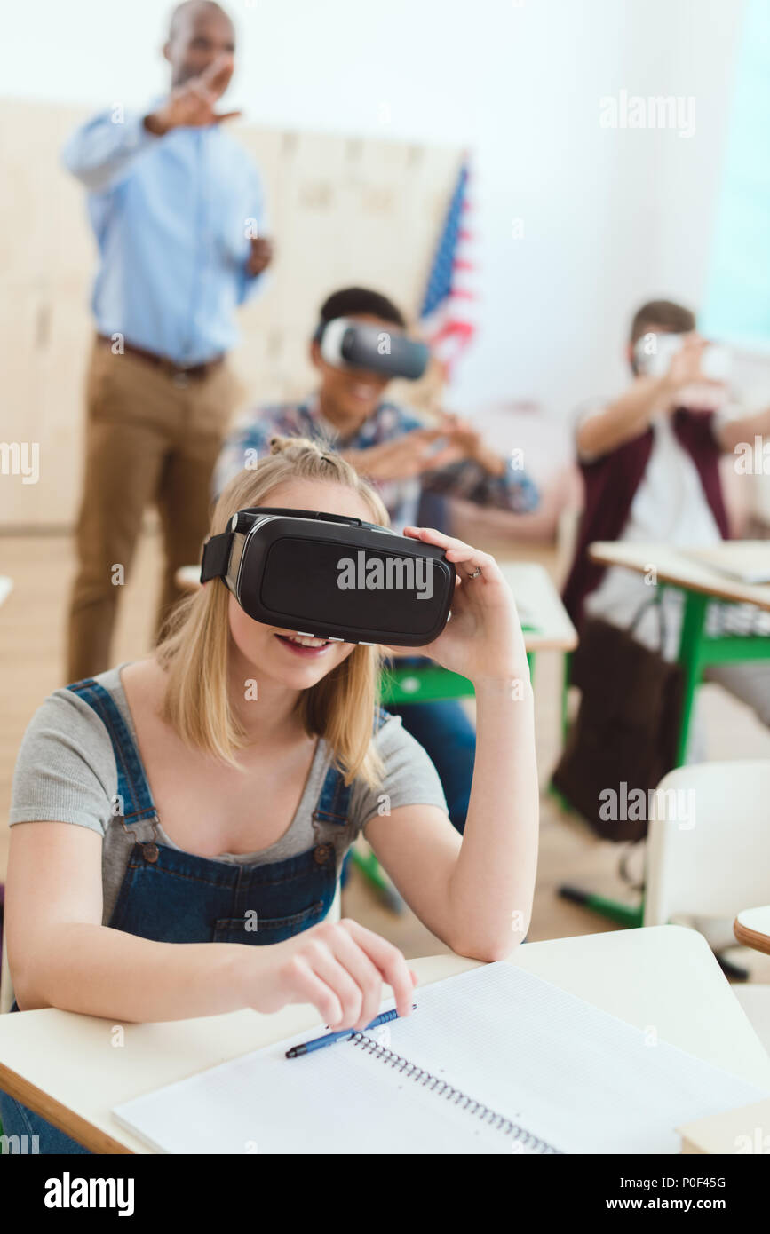 Teenage schoolgirl using virtual reality headset with male classmates ...