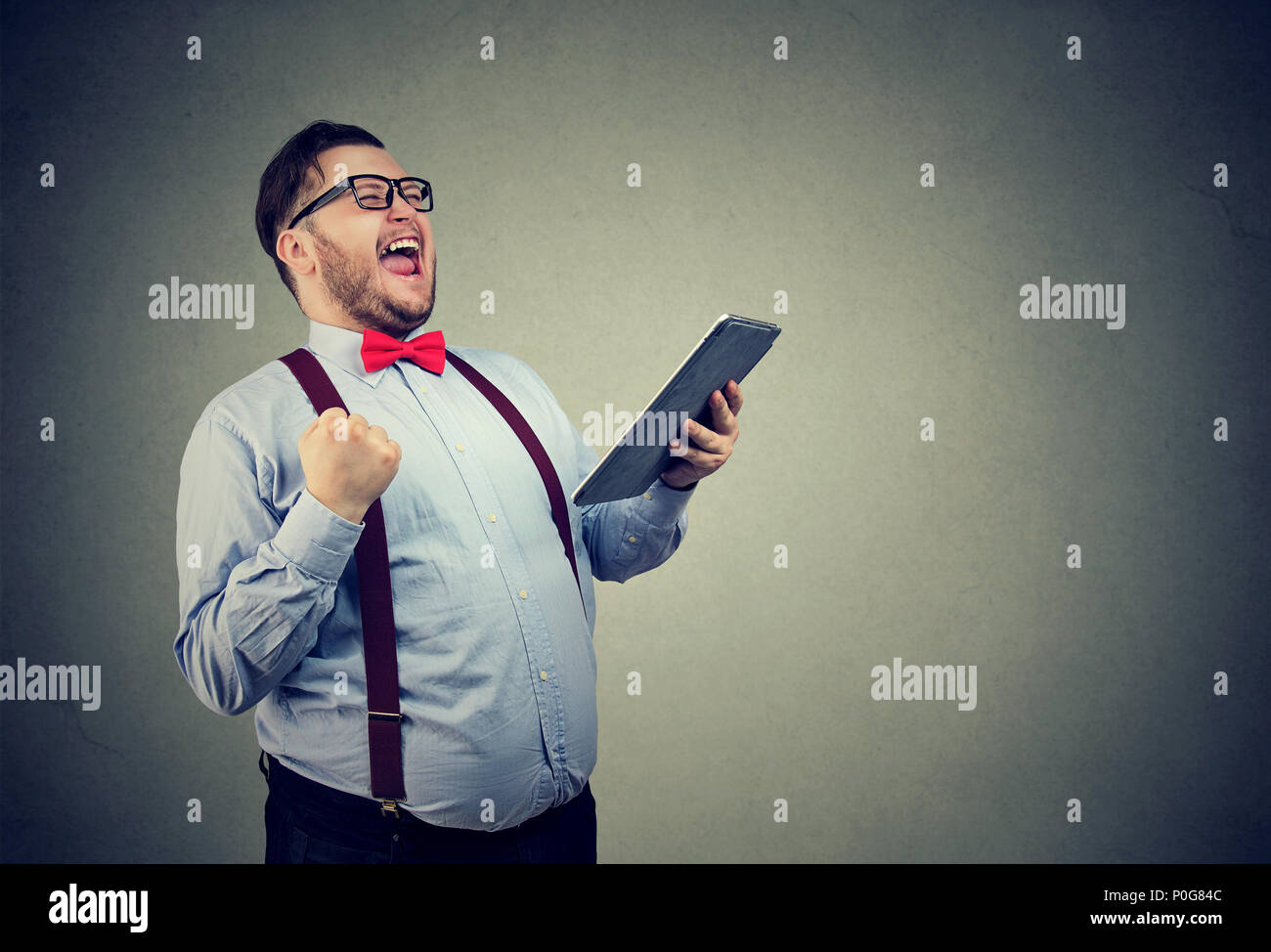 Trendy overweight business man in bow tie and glasses holding tablet and looking super excited with good news Stock Photo
