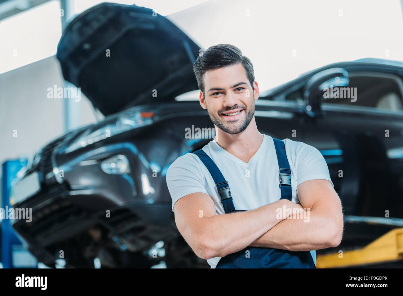 Smiling young automechanic standing at a repairshop in front of a lifted up car. Stock Photo