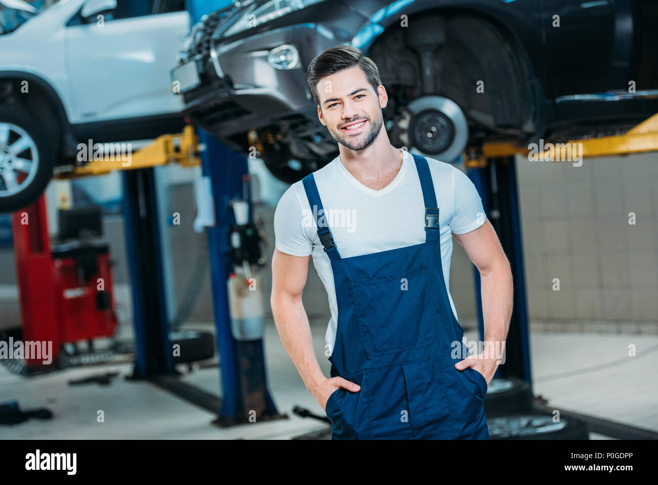 Smiling young automechanic standing at a repairshop in front of a lifted up car. Stock Photo