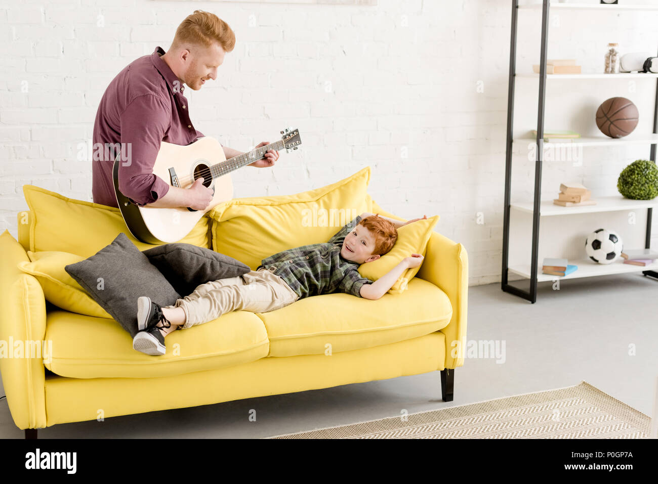 happy father playing guitar and cute smiling little son lying on couch Stock Photo