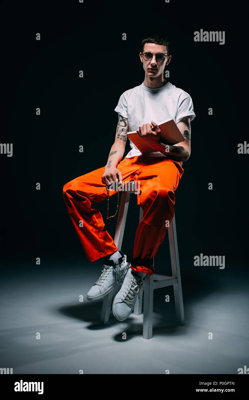 Serious Man In Orange Uniform With Cuffs On Legs Reading Bible And Sitting  On Stool On Dark Background Stock Photo - Alamy