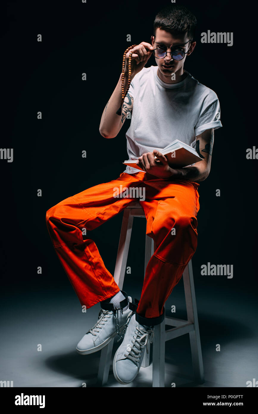 Man In Orange Uniform With Cuffs On Legs Reading Bible While Sitting On  Stool On Dark Background Stock Photo - Alamy