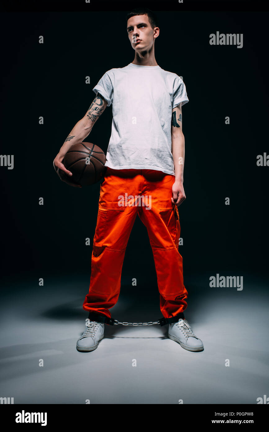 Man wearing prison uniform and cuffs holding basketball ball on dark background Stock Photo