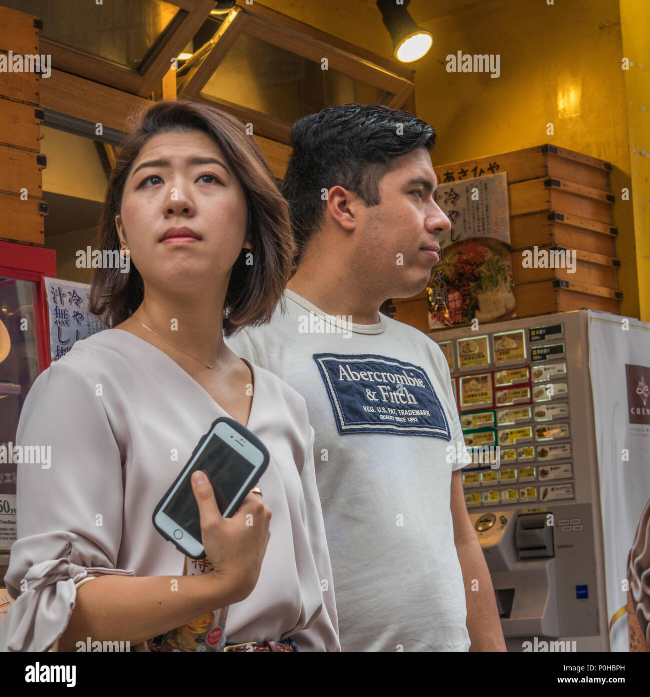 Standing together, looking away. Attractive young couple, asian woman, western man,   white shirts, brown skin, black hair. On the street, Shinjuku, T Stock Photo