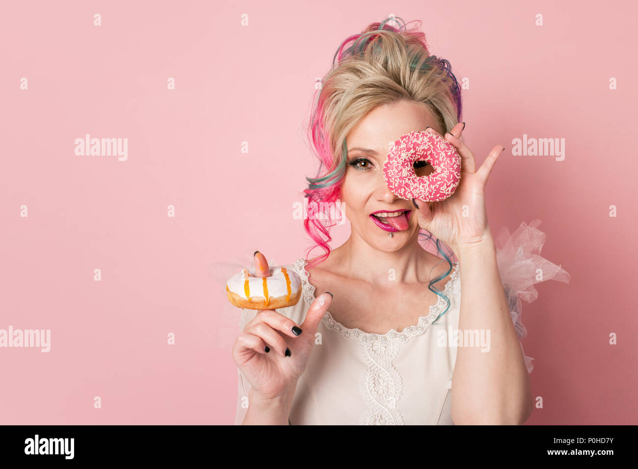 Stylish and beautiful woman with colored hair. Two donuts as glasses, funny grimace. Pink background Stock Photo