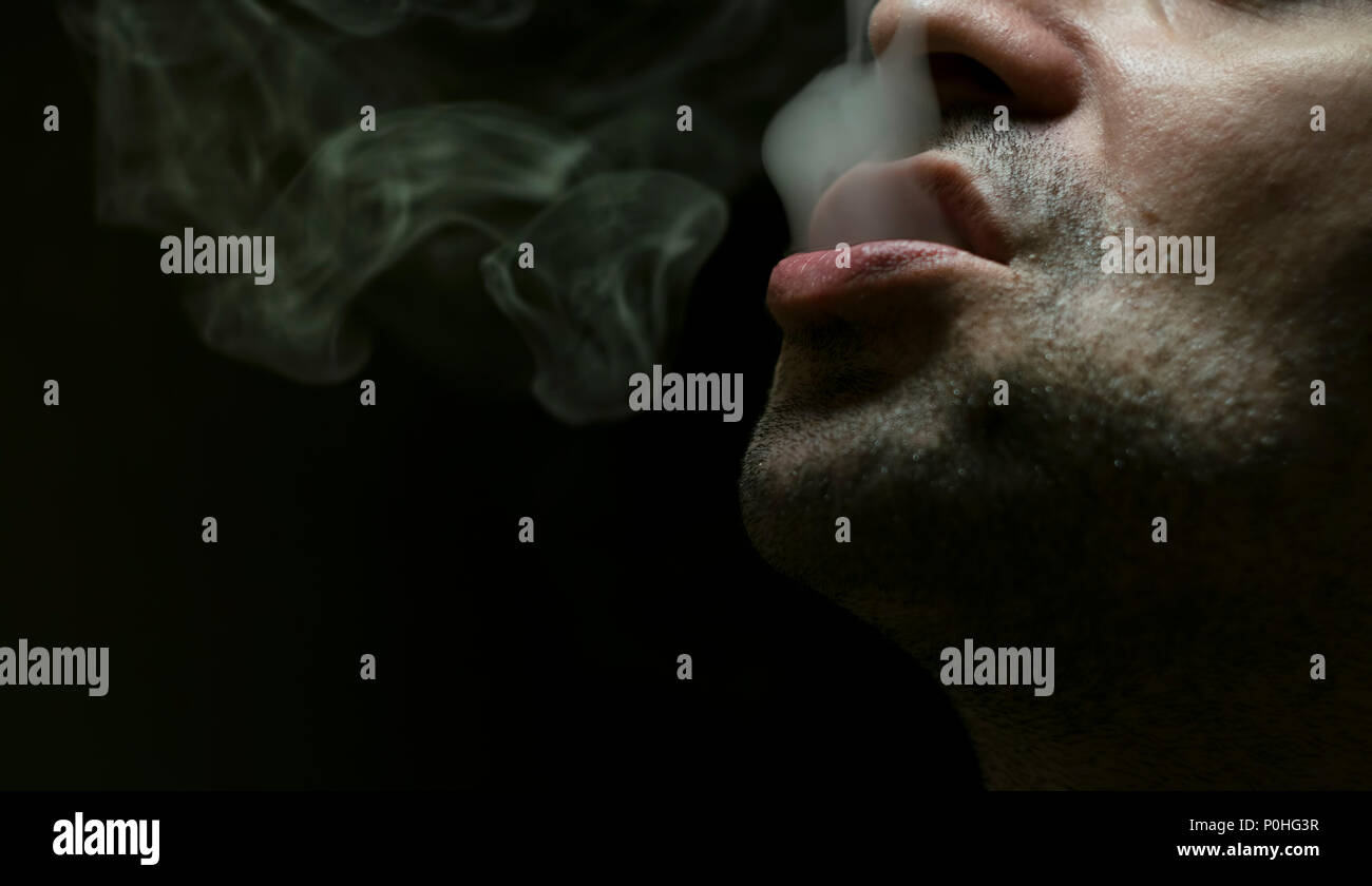 Close up shot of a man exhaling cigarette smoke on black background with dramatic light. Stock Photo