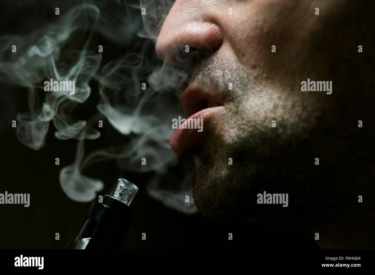 Close up shot of a man exhaling e-cigarette smoke on black background with dramatic light. Stock Photo