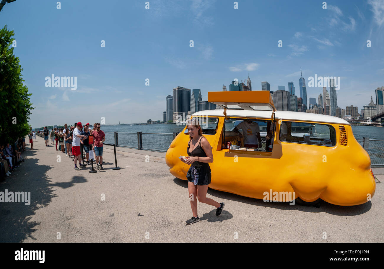 Brooklyn, USA. 9th June 2018. With the New York skyline behind them visitors to Brooklyn Bridge Park in New York delight in artist Erwin Wurm's 'Hot Dog Bus' serving free hot dogs to any and all, seen on opening day, Saturday, June 9, 2018. The Austrian artist modified a vintage Volkswagen Microbus into a bulbous bright yellow food truck, redefining the dichotomy between commerce and sculpture, albeit serving free iconic New York street food. Credit: Richard Levine/Alamy Live News Stock Photo