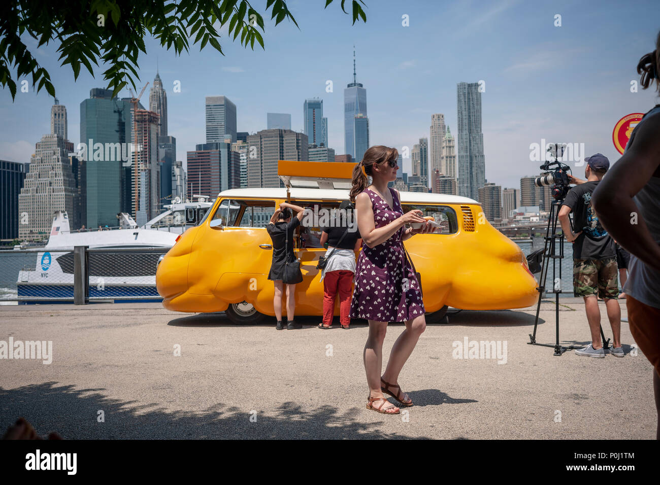Brooklyn, USA. 9th June 2018. With the New York skyline behind them visitors to Brooklyn Bridge Park in New York delight in artist Erwin Wurm's 'Hot Dog Bus' serving free hot dogs to any and all, seen on opening day, Saturday, June 9, 2018. The Austrian artist modified a vintage Volkswagen Microbus into a bulbous bright yellow food truck, redefining the dichotomy between commerce and sculpture, albeit serving free iconic New York street food. The truck was previously the 'Curry Bus' serving wursts in Europe. Credit: Richard Levine/Alamy Live News Stock Photo