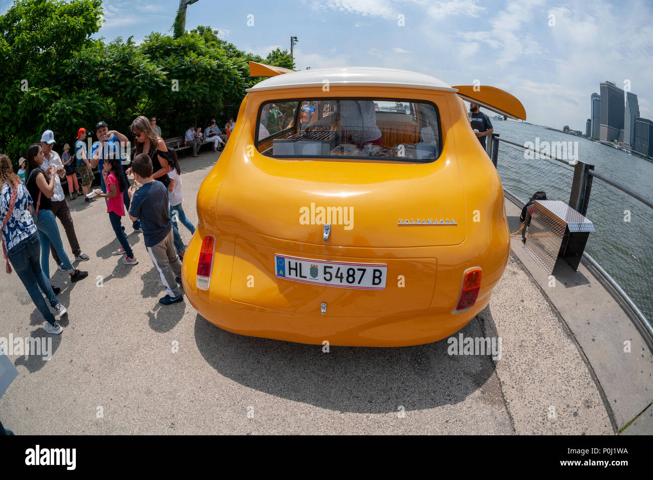 Brooklyn, USA. 9th June 2018. With the New York skyline behind them visitors to Brooklyn Bridge Park in New York delight in artist Erwin Wurm's 'Hot Dog Bus' serving free hot dogs to any and all, seen on opening day, Saturday, June 9, 2018. The Austrian artist modified a vintage Volkswagen Microbus into a bulbous bright yellow food truck, redefining the dichotomy between commerce and sculpture, albeit serving free iconic New York street food. The truck was previously the 'Curry Bus' serving wursts in Europe. Credit: Richard Levine/Alamy Live News Stock Photo