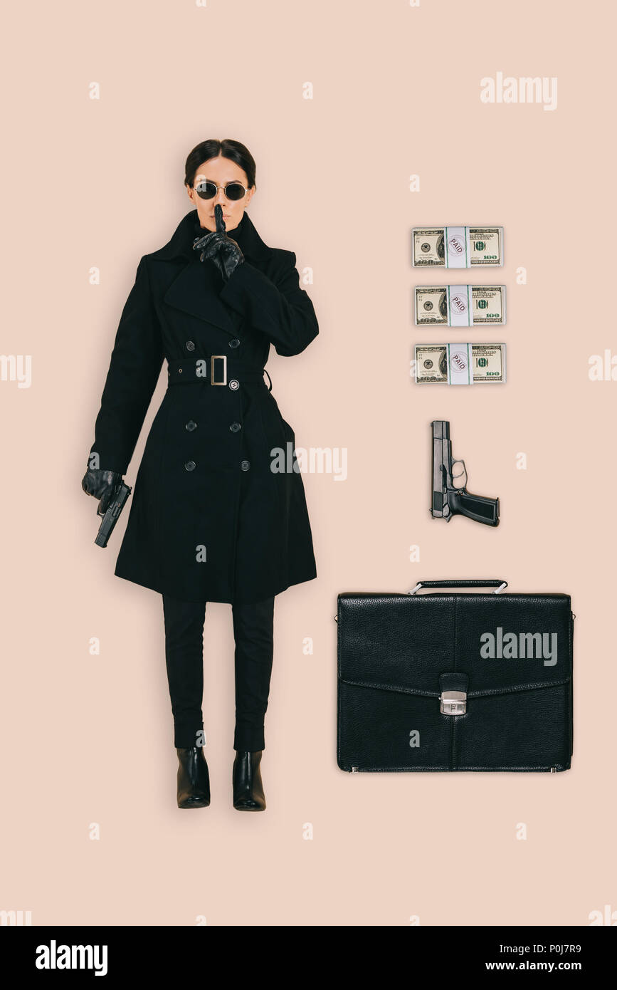 top view of female killer in sunglasses doing shushing gesture with briefcase, handguns and cash isolated on pink background Stock Photo