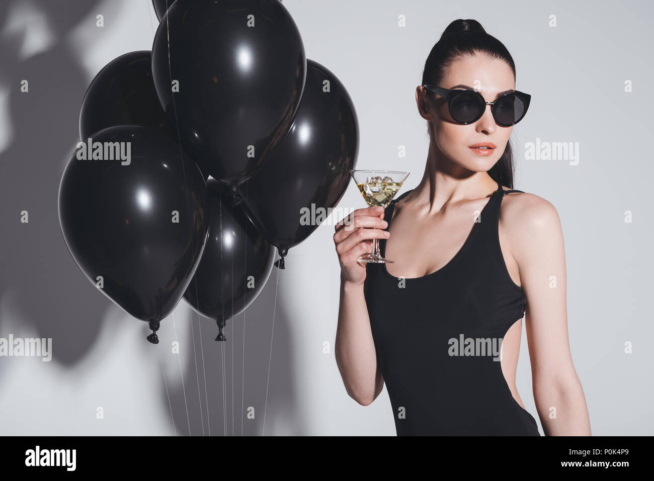 Gorgeous young woman in sunglasses and swimsuit drinking martini while standing in studio with black balloons Stock Photo