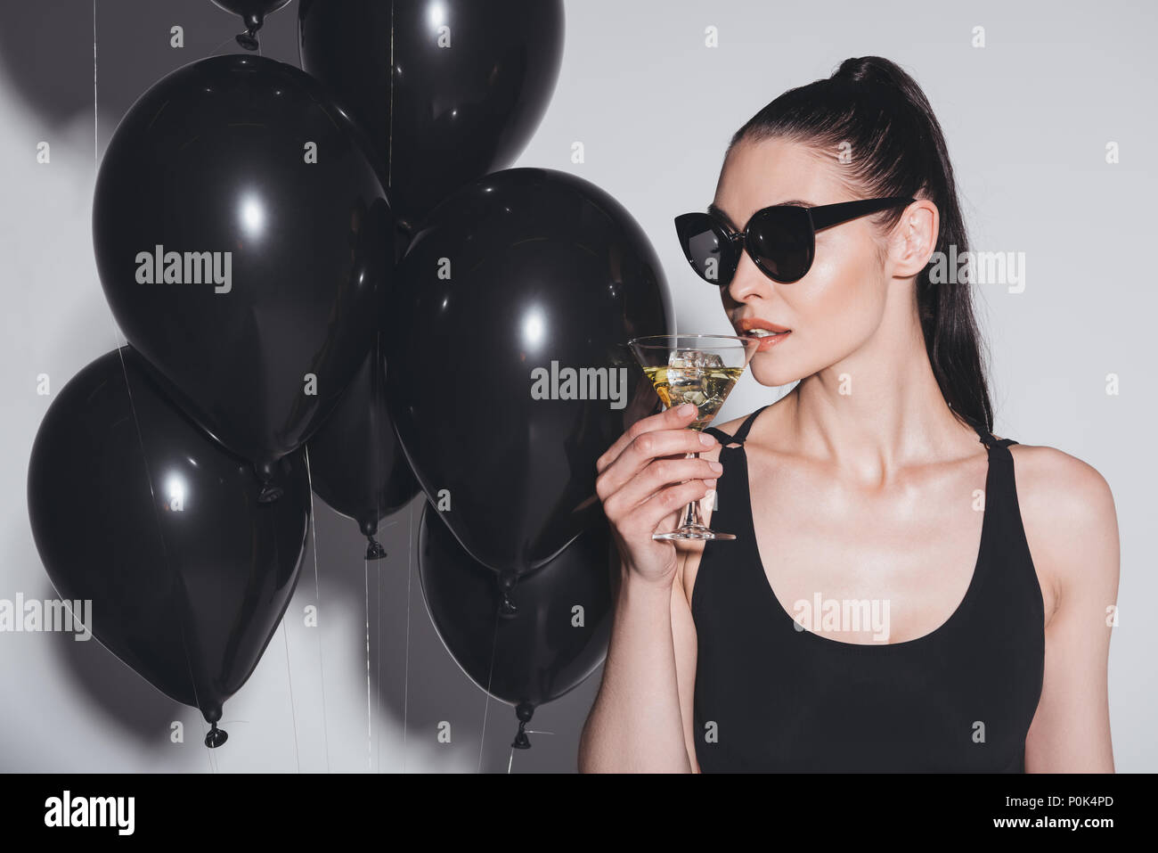 Gorgeous young woman in sunglasses and leotard drinking martini while standing in studio with black balloons Stock Photo