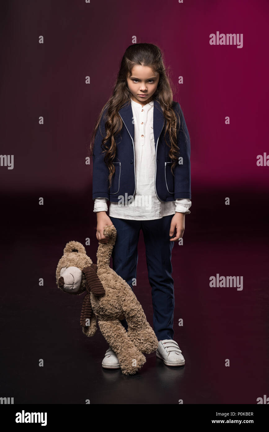 angry kid standing and holding teddy bear on burgundy Stock Photo