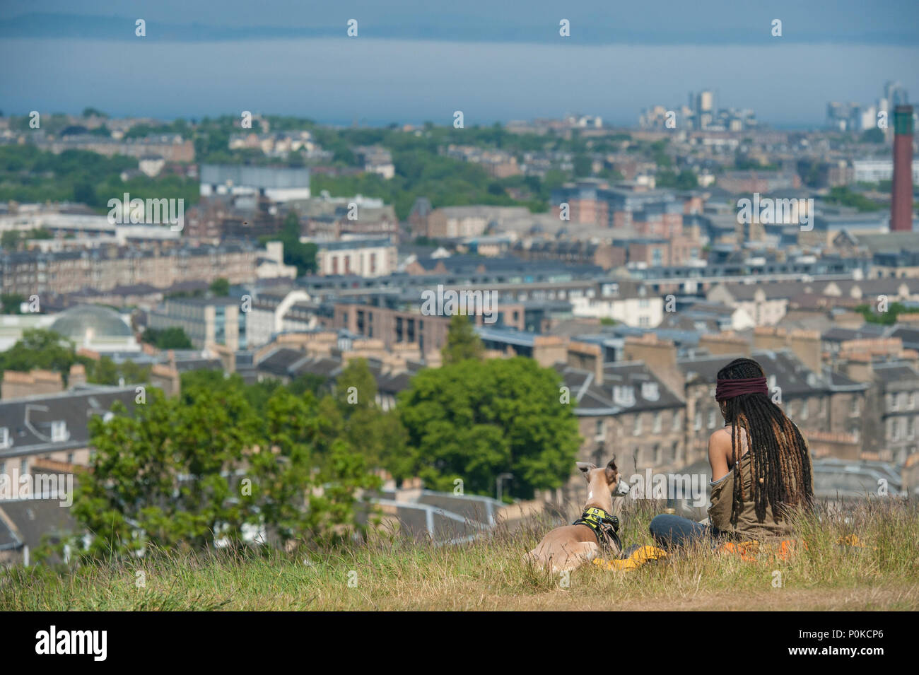 A very cool, bohemian girl sits in the long grass on Carlton Hill on a sunny day with Edinburgh below her and her dog Stock Photo