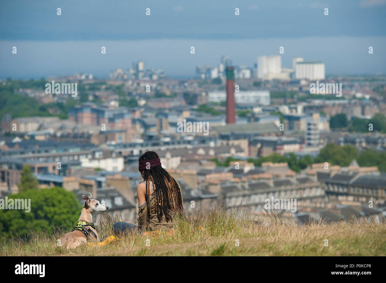A very cool, bohemian girl sits in the long grass on Carlton Hill on a sunny day with Edinburgh below her and her dog Stock Photo