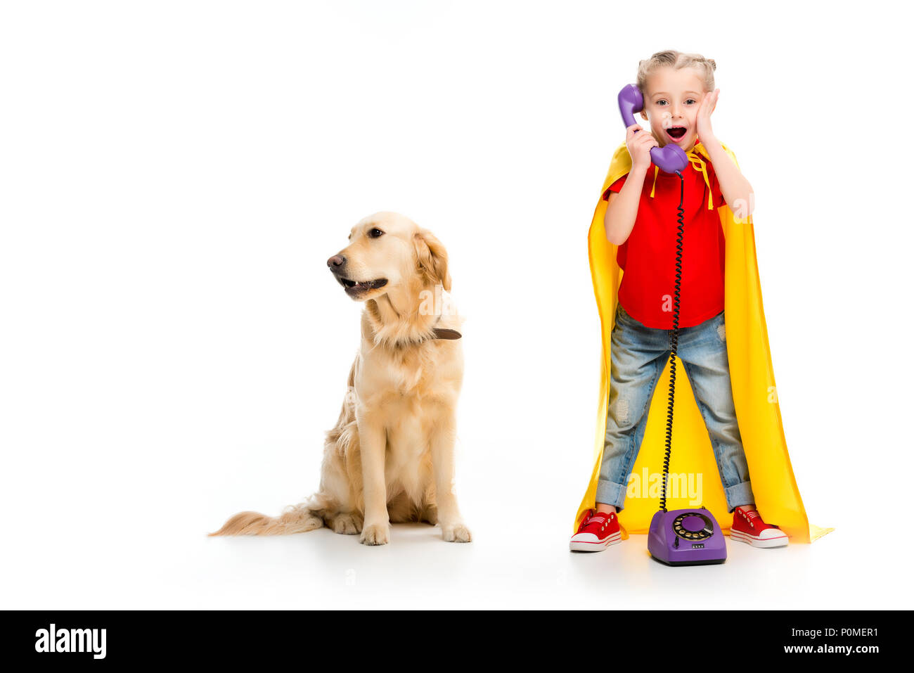 Shocked little supergirl wearing yellow cape and talking on phone with standing dog beside isolated on white Stock Photo