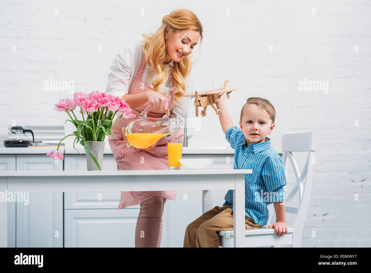 Mother pouring orange juice for little son while he playing with wooden airplane Stock Photo