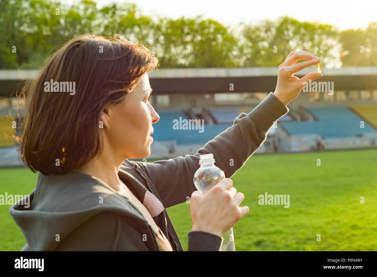 Outdoor portrait of mature woman taking vitamin E capsule pill of cod liver oil, at the stadium. Sport, healthy lifestyle and nutrition, healthcare co Stock Photo