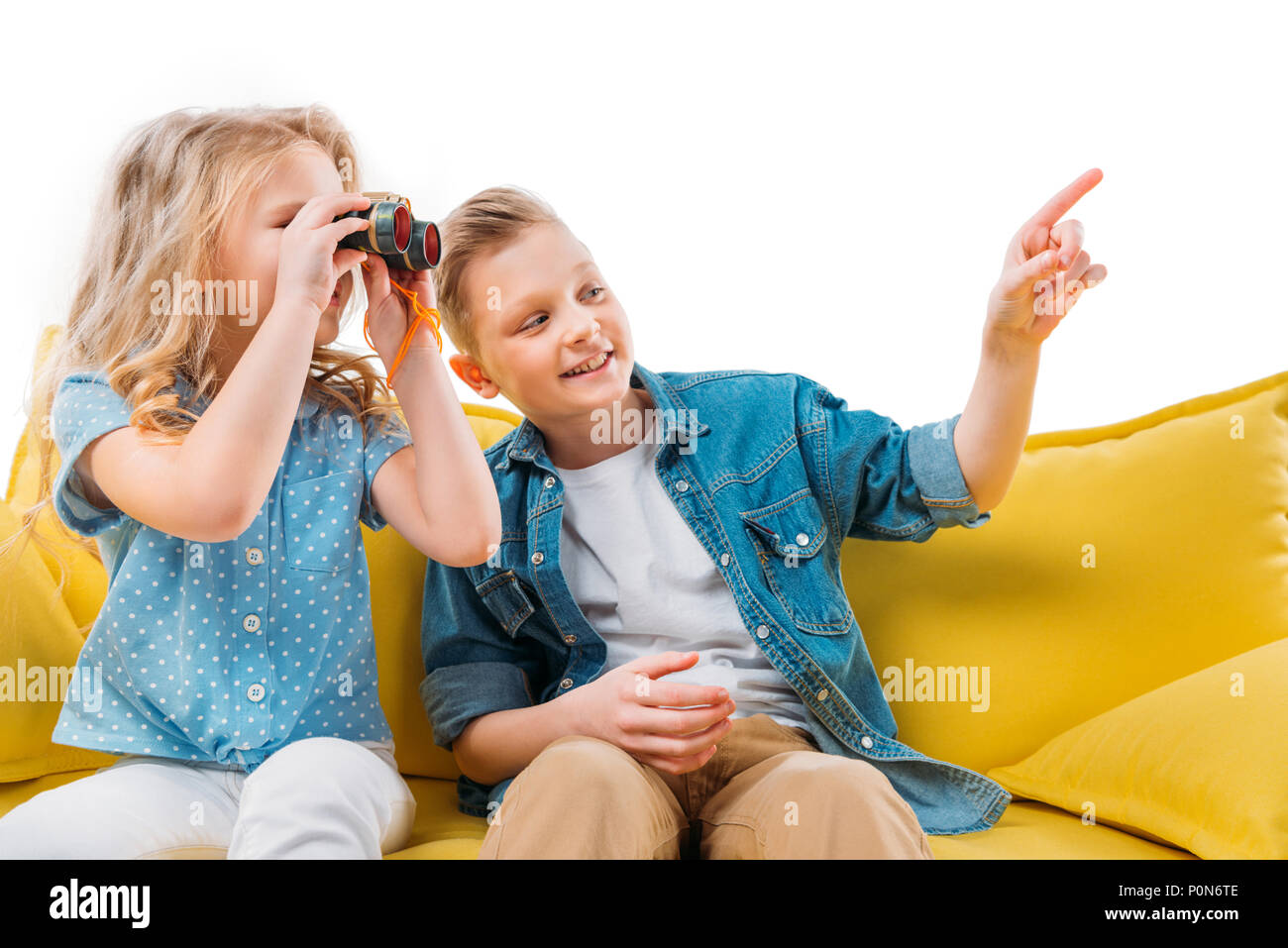 boy pointing while sister looking at binoculars, sitting on yellow sofa Stock Photo