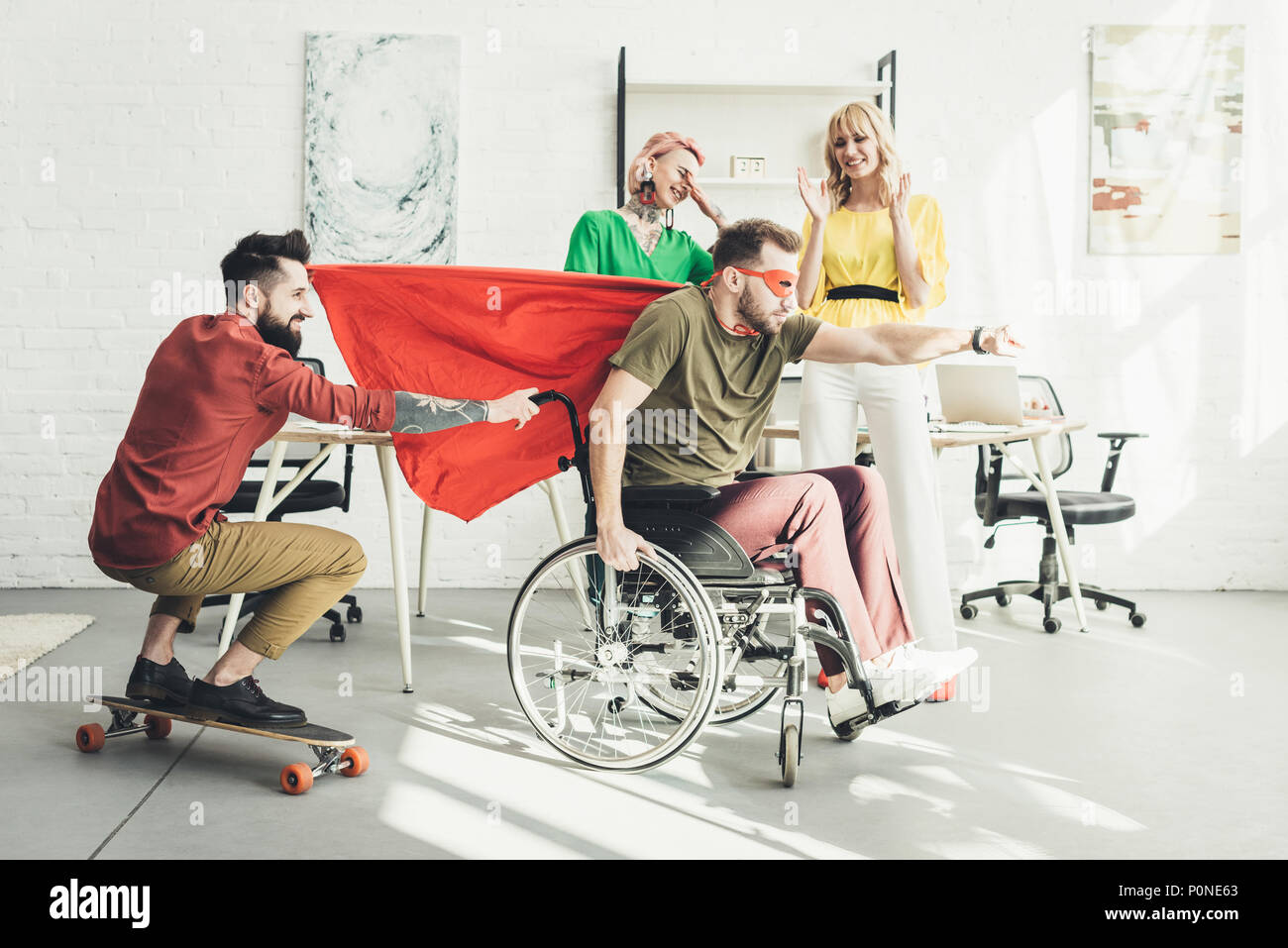 disabled businessman in superhero costume with colleague on skateboard and businesswomen behind in office Stock Photo