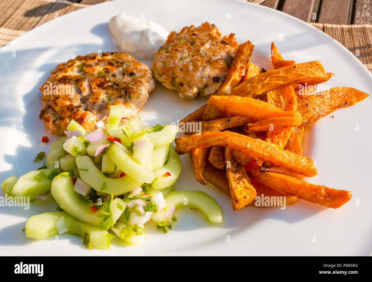 Close up of white dinner plate with Southeast Asian food served in sunshine. Salmon fishcakes, cucumber salad and sweet potato chips Stock Photo