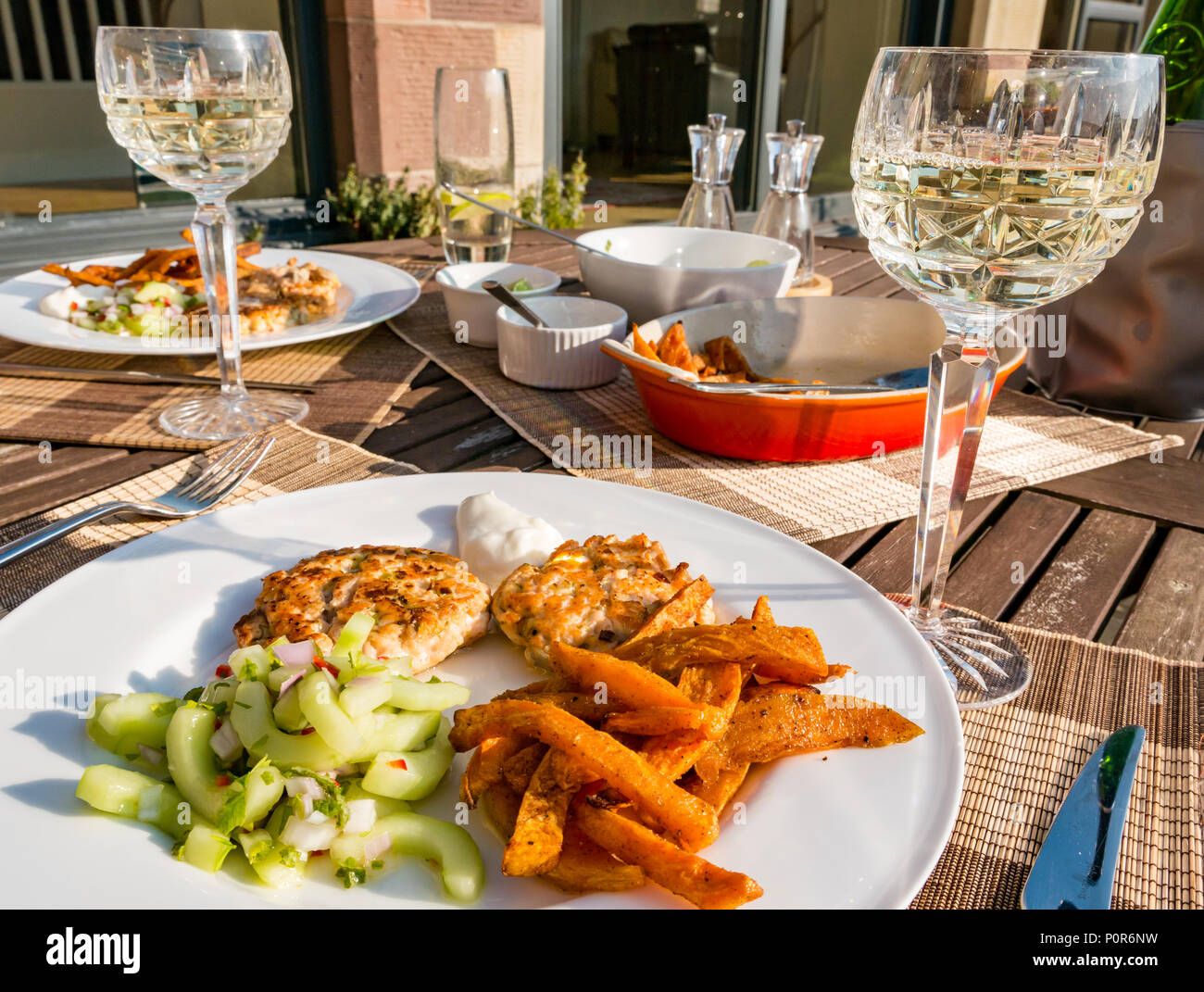 Southeast Asian food served on outdoor patio table in sunshine with crystal wine glasses filled with white wine. Salmon fishcakes, cucumber salad and sweet potato chips Stock Photo