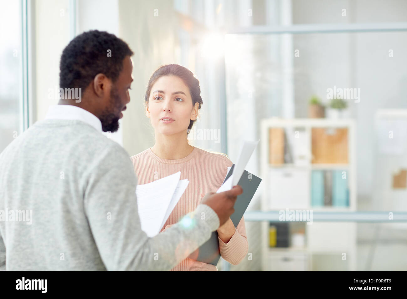 Bearded African American entrepreneur studying statistic data with young subordinate while standing at modern open plan office, lens flare Stock Photo