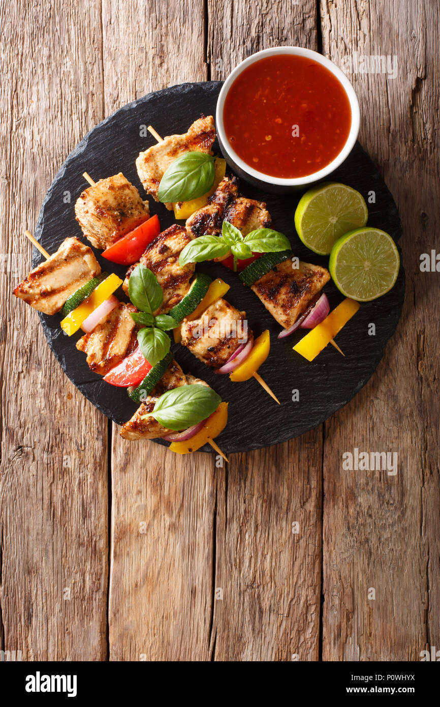 Hot grilled chicken kebab skewers with colorful vegetables with sauce, basil and lime closeup on the table. Vertical top view from above Stock Photo