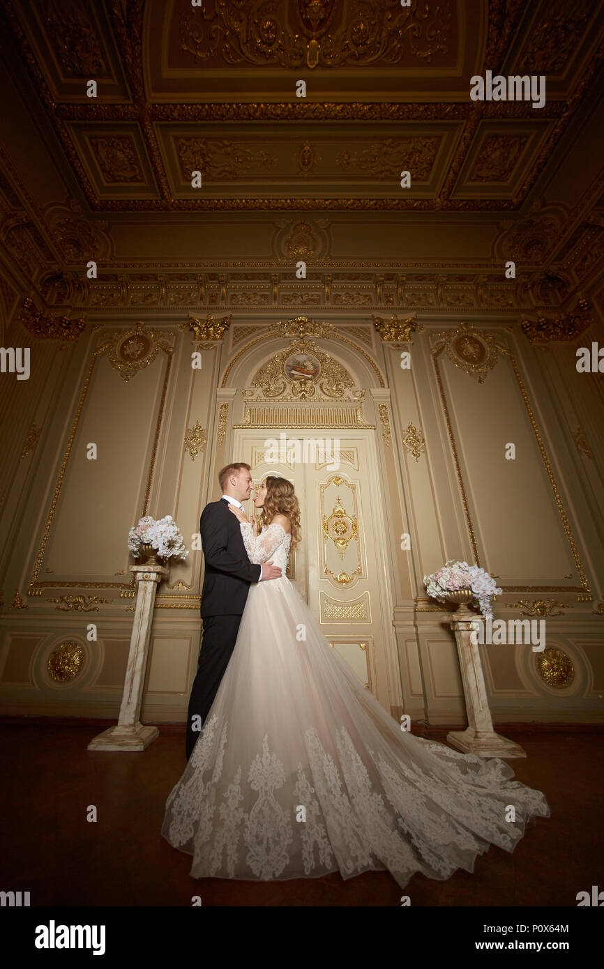 Luxury wedding couple in love. Beautiful bride in white dress with brides  bouquet and handsome groom in black suit standing in baroque interior and  embracing each other Stock Photo - Alamy