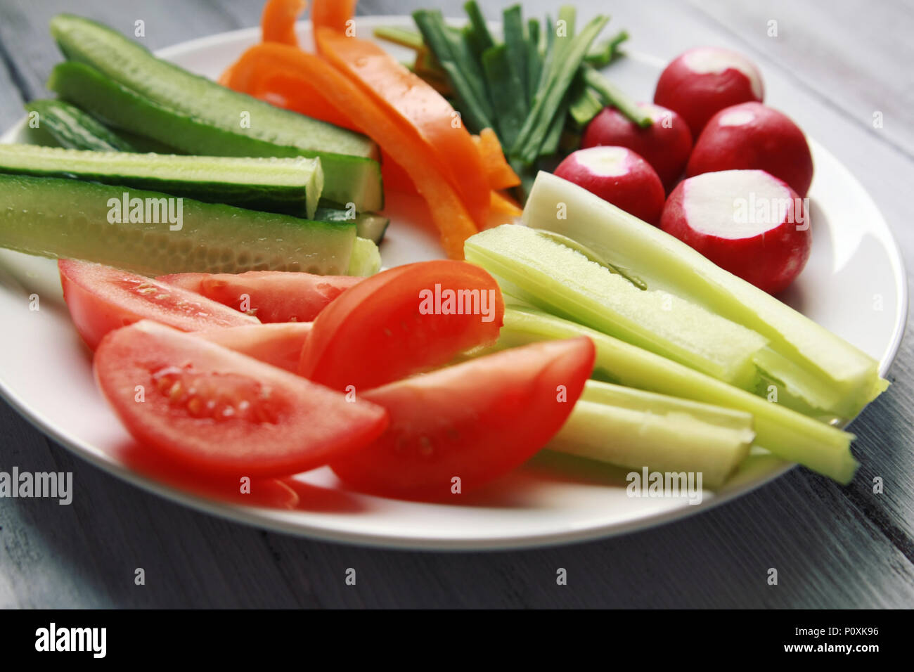 White plate with vegetables for a vegetarian salad. Radishes, tomatoes, celery, bell pepper, onion and cucumber. White wooden kitchen table. Close up. Stock Photo